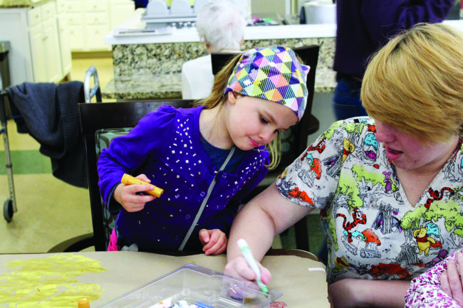 Elsa Cheney (left) and Ashley, a care partner at Jill’s House, color on paper-covered tables during a field trip to visit the elder residents who live on the upper level of Jill’s House