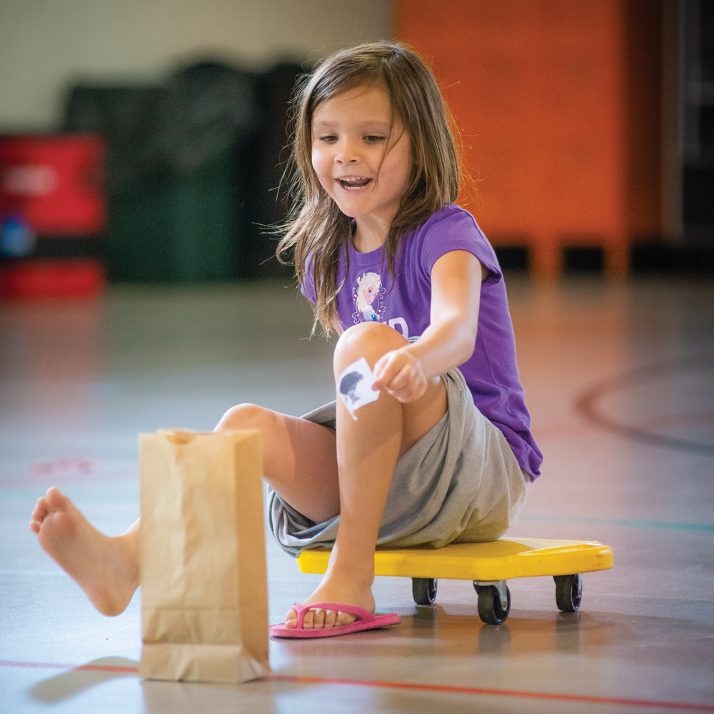 Six-year-old Riley slides across the floor to drop a piece of paper with a picture of a food into a paper bag corresponding to its food group during Food Group Relay.