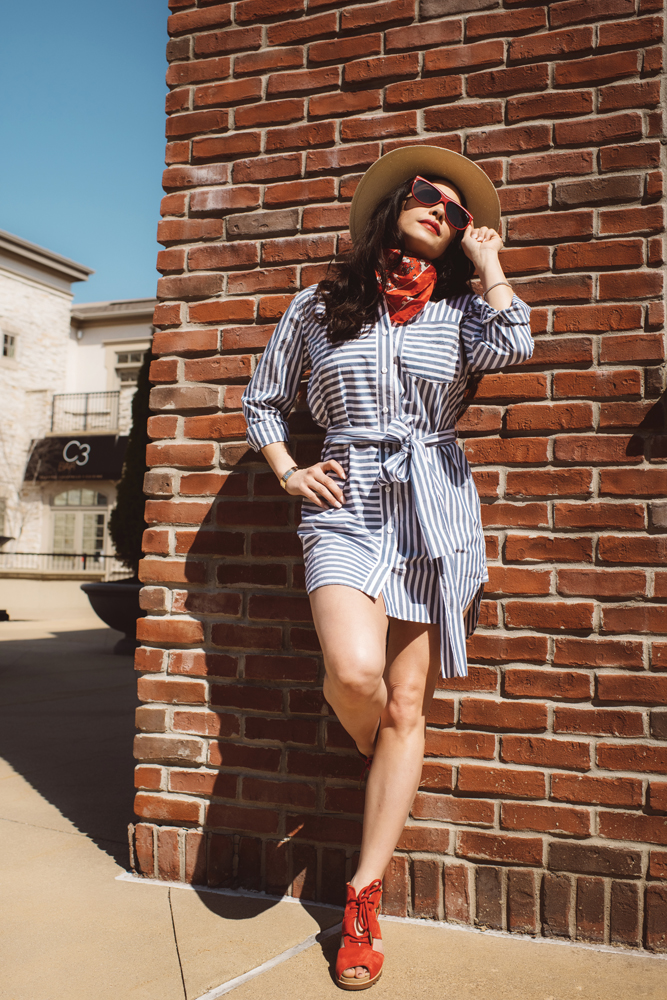 Morgan Miller models a stylish ensemble featuring a striped cotton dress by Current/Elliott, a straw fedora by Rag & Bone, sunglasses by Quay, a floral scarf by Rag & Bone, and suede wedges by Sorel.