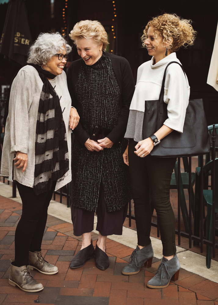 Diane Harrison West wears an asymmetric tunic by Et’Lois and M Rena leggings with a silk burnout scarf by Blue Pacific, Barbara Weiskopf wears a textured high-collared coat from Chalet, and Samantha Eibling wears a Chalet cowl-neck sweater with a vegan leather bracelet and handbag from Joy.